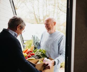 A senior man is delivering a box full of groceries to his neighbour / Un homme âgé livre un carton rempli de provisions à son voisin